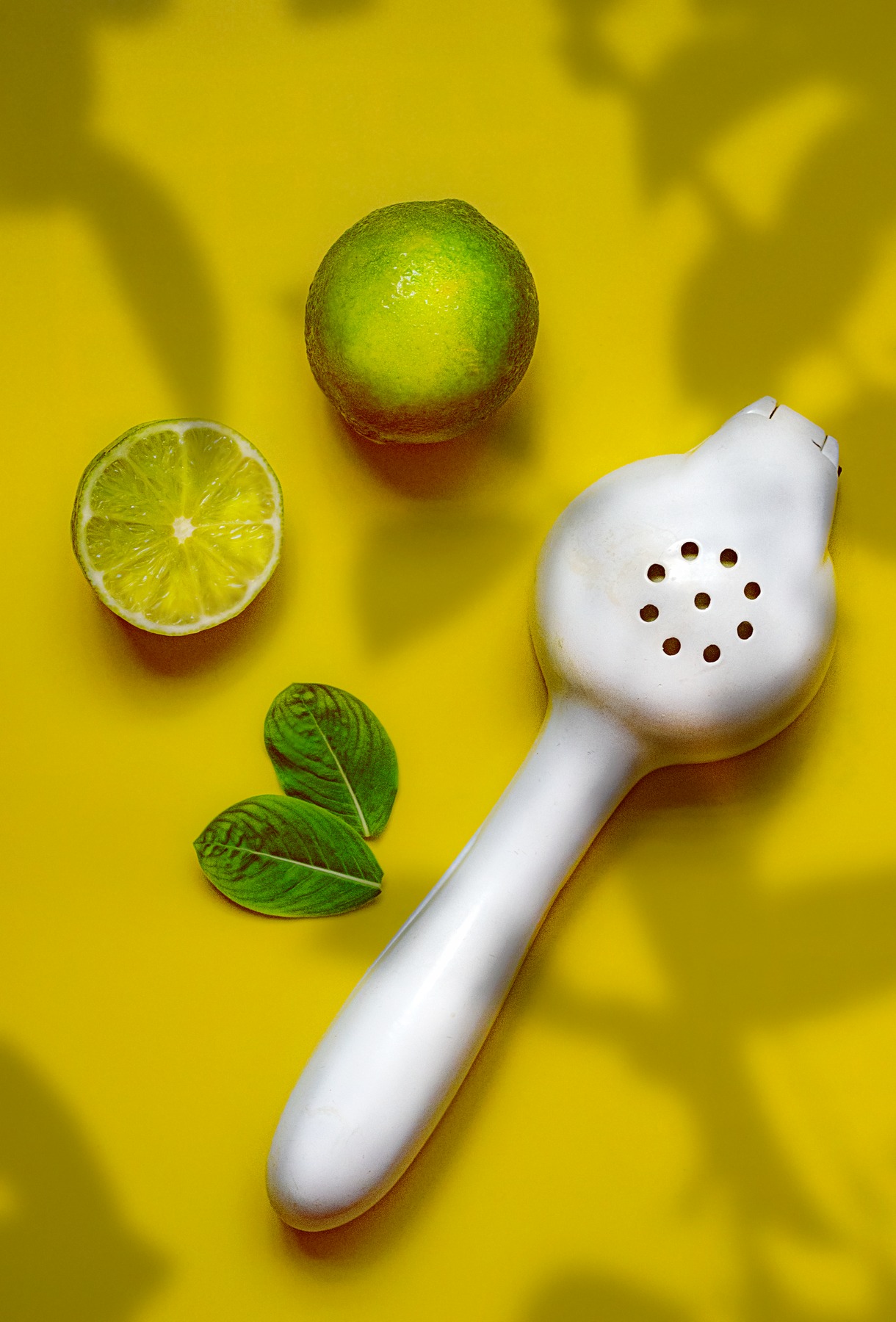 A whole lime, a cut lime and a white citrus press on a yellow surface.