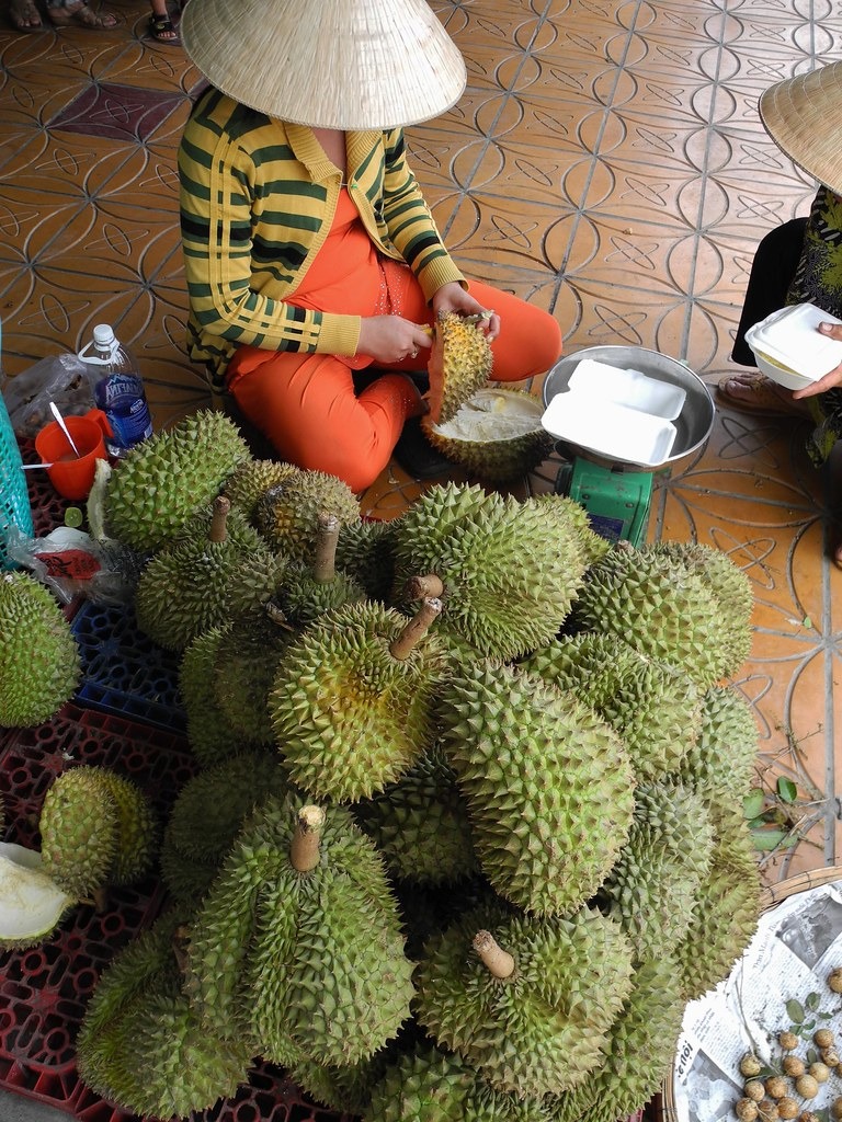 A market vendor in a conical hat opening durian beside a pile of whole fruit.