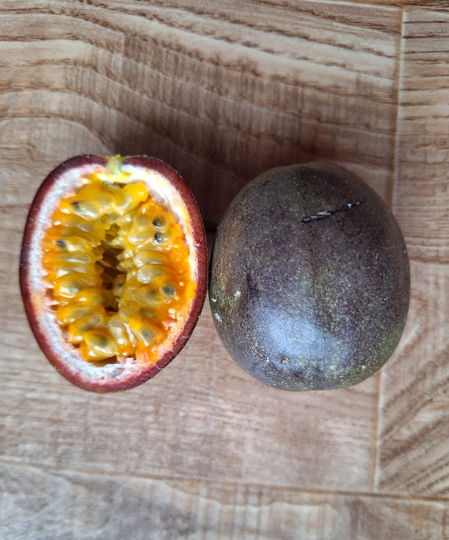 A whole purple passion fruit beside a cut half on a wooden surface.